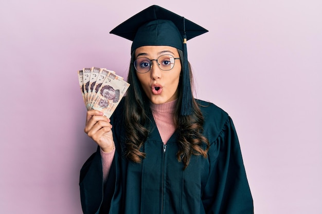 Joven hispana con uniforme de graduación sosteniendo billetes de pesos mexicanos asustada y sorprendida con la boca abierta por sorpresa, cara de incredulidad