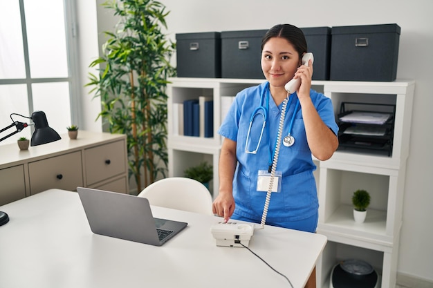 Joven hispana con uniforme médico hablando por teléfono en la clínica