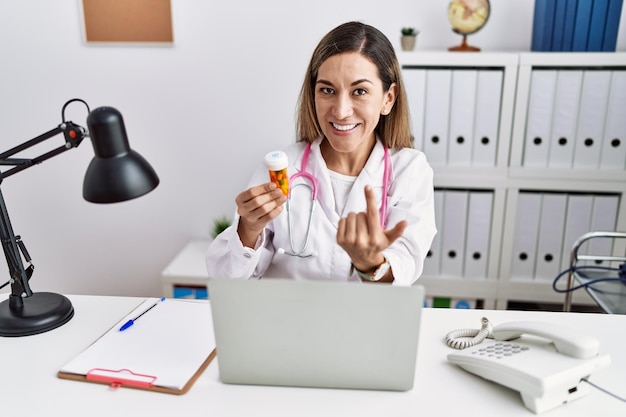 Joven hispana con uniforme médico sosteniendo pastillas en la clínica llamando a venir aquí gesto con la mano invitando a dar la bienvenida feliz y sonriente