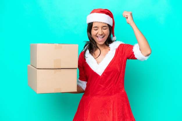 Foto joven hispana vestida como mamá noel sosteniendo una caja de dibujos animados aislada de fondo azul celebrando una victoria