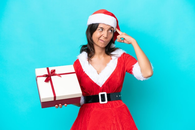 Joven hispana vestida como mamá noel sosteniendo un regalo aislado de fondo azul con dudas y pensando