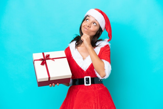 Joven hispana vestida como mamá noel sosteniendo un regalo aislado de fondo azul y mirando hacia arriba