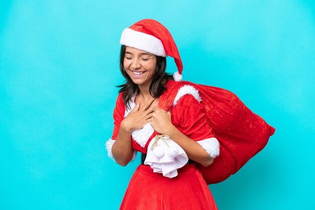 Joven hispana vestida como mamá noel sosteniendo un saco aislado de fondo azul sonriendo mucho