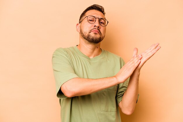 Joven hispano aislado de fondo beige sintiéndose enérgico y cómodo frotándose las manos confiado