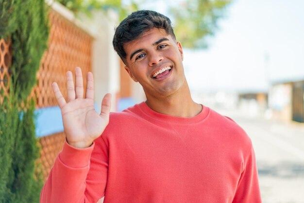 Foto joven hispano al aire libre saludando con la mano con expresión feliz