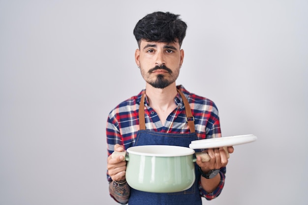 Joven hispano con barba con delantal sosteniendo una olla relajada con una expresión seria en la cara simple y natural mirando a la cámara