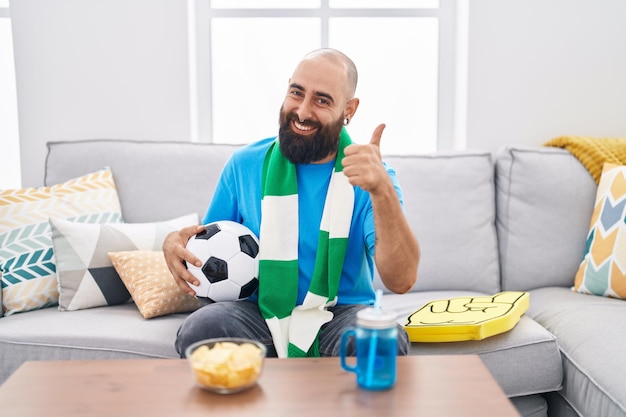 Joven hispano con barba y tatuajes hooligan de fútbol sosteniendo el equipo de apoyo de la pelota sonriendo feliz y positivo, pulgar arriba haciendo excelente y signo de aprobación