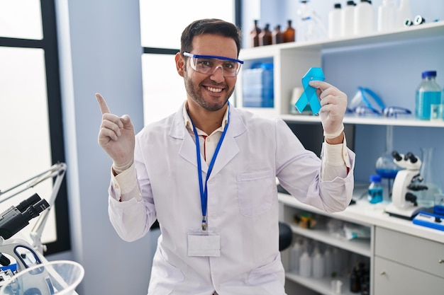 Joven hispano con barba trabajando en un laboratorio científico sosteniendo una cinta azul sonriendo feliz señalando con la mano y el dedo a un lado