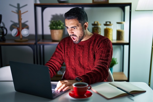 Joven hispano con barba usando computadora portátil por la noche en casa con cara de shock mirando escéptico y sarcástico sorprendido con la boca abierta