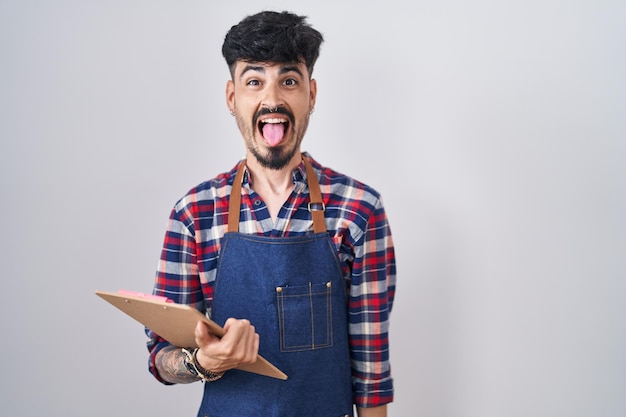 Joven hispano con barba usando delantal de camarero sosteniendo el portapapeles sacando la lengua feliz con expresión divertida concepto de emoción