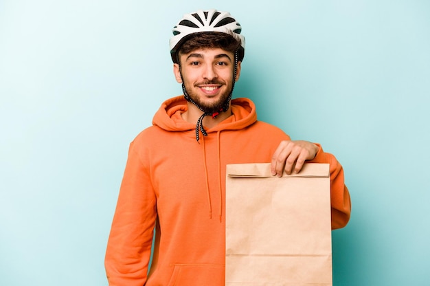 Joven hispano con una bicicleta con casco sosteniendo una comida para llevar aislada de fondo azul feliz sonriendo y alegre