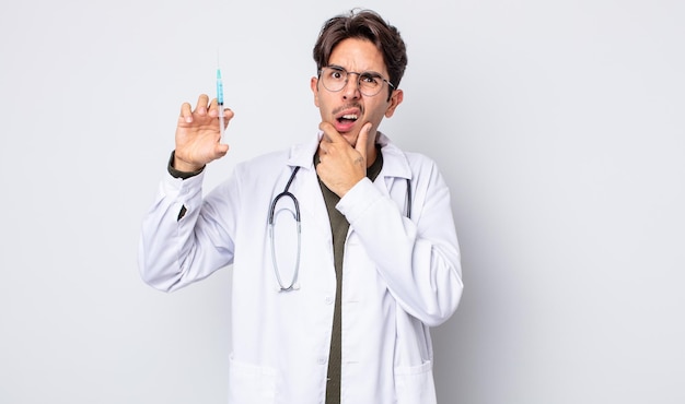 Joven hispano con la boca y los ojos bien abiertos y la mano en el concepto de jeringa del médico de la barbilla