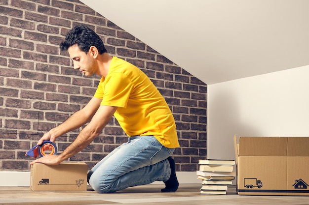 Joven hispano de cabello oscuro con camiseta amarilla y jeans azules que se muda de casa cerrando cajas en el ático de su apartamento