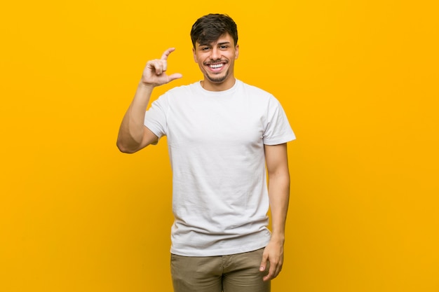 Joven hispano casual sosteniendo algo poco con los dedos índices, sonriente y confiado.