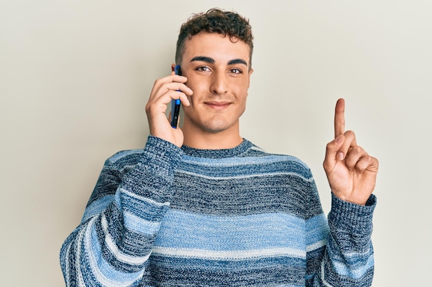 Joven hispano conversando hablando en el teléfono inteligente sonriendo con una idea o pregunta señalando con el dedo con la cara feliz número uno