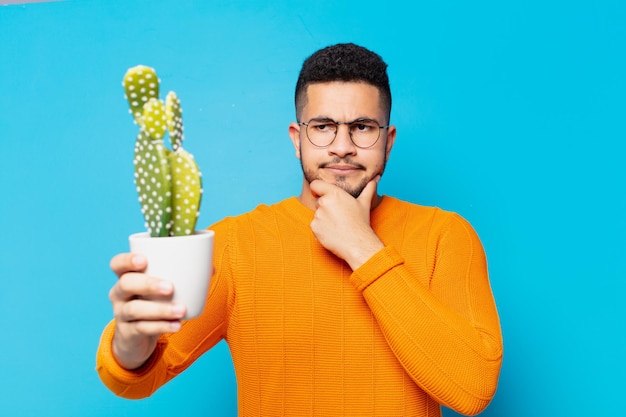 Joven hispano dudando o expresión incierta y sosteniendo un cactus