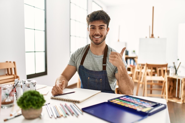 Joven hispano en el estudio de arte sonriendo feliz y positivo, pulgar arriba haciendo excelente y señal de aprobación