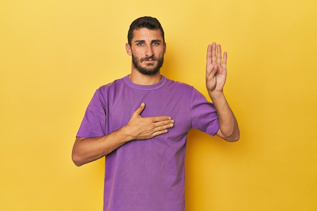 Foto joven hispano de fondo amarillo prestando juramento poniendo la mano en el pecho