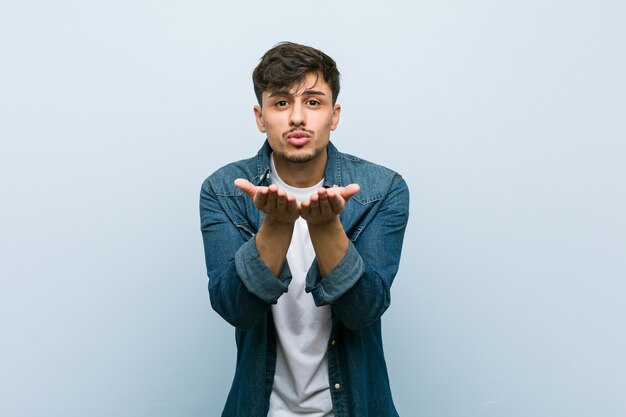 Joven hispano fresco doblando los labios y sosteniendo las palmas de las manos para enviar un beso al aire.