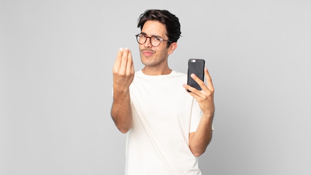 Foto joven hispano haciendo capice o gesto de dinero, diciéndole que pague y sosteniendo un teléfono inteligente