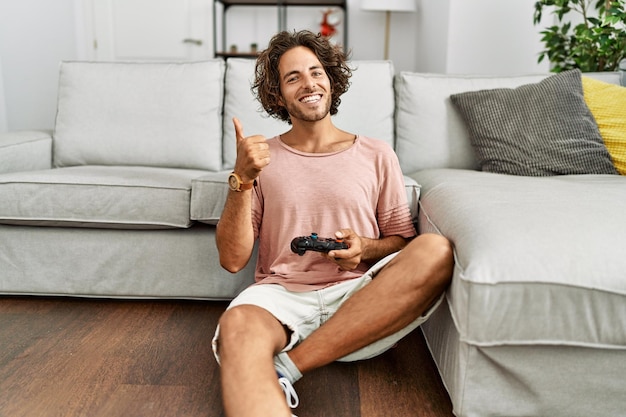 Joven hispano jugando videojuegos sosteniendo el controlador en casa sonriendo feliz y positivo, pulgar arriba haciendo excelente y señal de aprobación