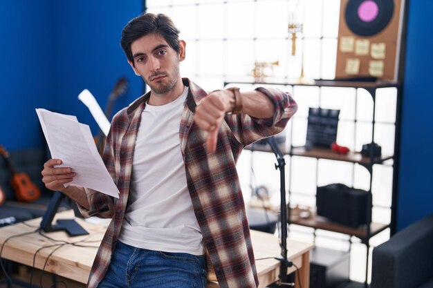 Joven hispano leyendo partituras en el estudio de música con un signo negativo de cara enfadada que muestra disgusto con el concepto de rechazo de los pulgares hacia abajo