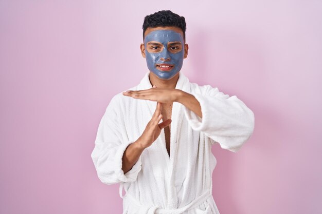 Joven hispano con mascarilla de belleza y bata de baño haciendo gesto de tiempo fuera con las manos, cara frustrada y seria