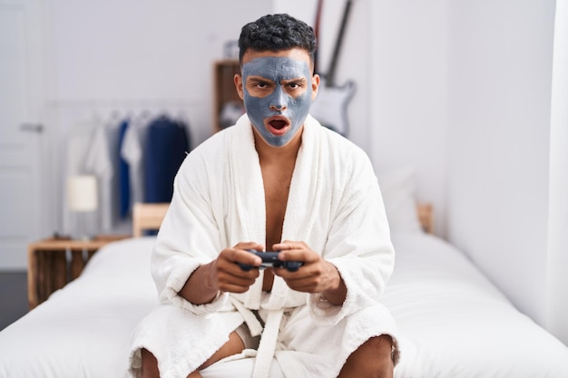 Joven hispano con mascarilla de belleza jugando videojuegos con cara de shock, luciendo escéptico y sarcástico, sorprendido con la boca abierta