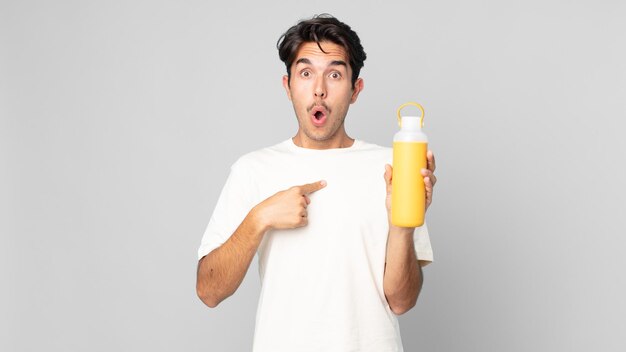 Joven hispano mirando consternado y sorprendido con la boca abierta, apuntando a sí mismo con un termo de café