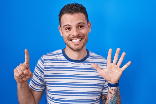 Joven hispano parado sobre un fondo azul que aparece y señala con los dedos número seis mientras sonríe confiado y feliz.