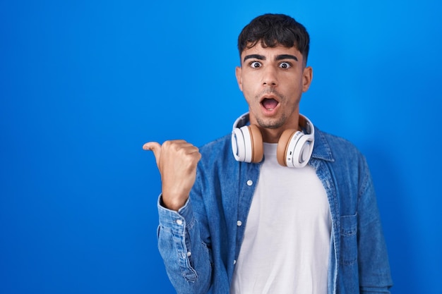 Joven hispano parado sobre fondo azul sorprendido señalando con el dedo de la mano hacia un lado, boca abierta expresión asombrada.