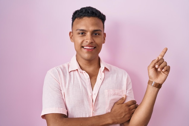 Joven hispano parado sobre un fondo rosado con una gran sonrisa en la cara, señalando con el dedo de la mano hacia un lado mirando la cámara.