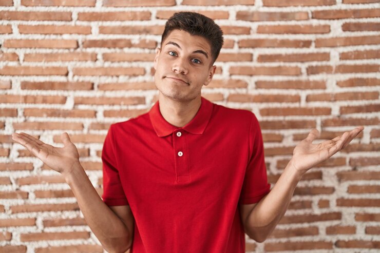 Foto joven hispano parado sobre una pared de ladrillos sin idea y expresión confusa con los brazos y las manos levantados. concepto de duda.