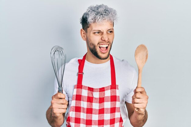 Joven hispano con el pelo teñido moderno con delantal de cocinero sosteniendo batidor y cuchara de panadero enojado y loco gritando frustrado y furioso, gritando de ira. rabia y concepto agresivo.