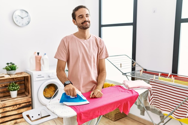 Joven hispano planchando ropa en casa mirando de lado a lado con una sonrisa en la cara, expresión natural. riendo confiado.