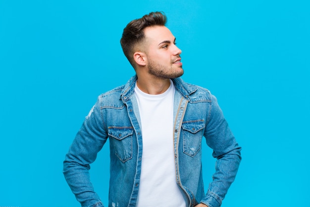 Joven hispano que parece feliz, alegre y confiado, sonriendo con orgullo y mirando al lado con ambas manos en las caderas contra la pared azul