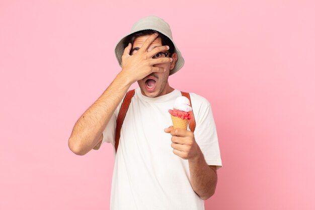 Joven hispano que parece sorprendido, asustado o aterrorizado, cubriendo la cara con la mano y sosteniendo un helado