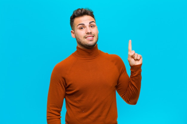 Joven hispano que se siente como un genio feliz y emocionado después de darse cuenta de una idea, levantando alegremente el dedo, ¡eureka! contra la pared azul