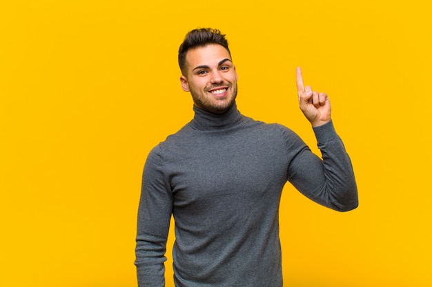 Joven hispano que se siente como un genio feliz y emocionado después de darse cuenta de una idea, levantando alegremente el dedo, ¡eureka! en la pared naranja