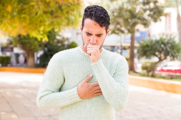 Foto joven hispano que se siente enfermo con dolor de garganta y síntomas de gripe tosiendo con la boca cubierta