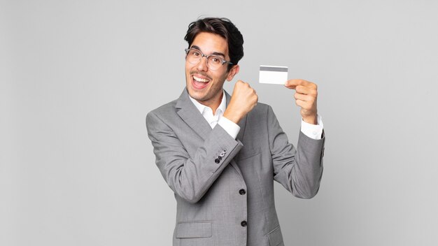 Joven hispano que se siente feliz y enfrenta un desafío o celebrando y sosteniendo una tarjeta de crédito