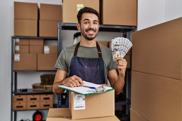 Joven hispano que trabaja en el comercio electrónico de pequeñas empresas con dinero con aspecto positivo y feliz de pie y sonriendo con una sonrisa segura mostrando los dientes