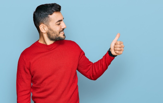 Joven hispano con ropa informal que se ve orgulloso sonriendo haciendo un gesto de aprobación hacia un lado