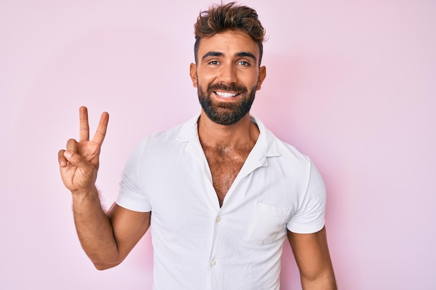 Joven hispano con ropa informal sonriendo con cara feliz guiñando un ojo a la cámara haciendo el signo de la victoria número dos