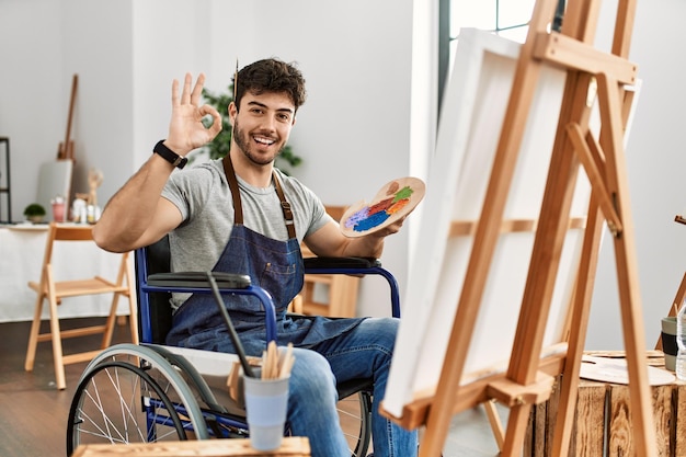 Joven hispano sentado en silla de ruedas pintando en un estudio de arte sonriendo positivamente haciendo el signo de ok con la mano y los dedos expresión exitosa