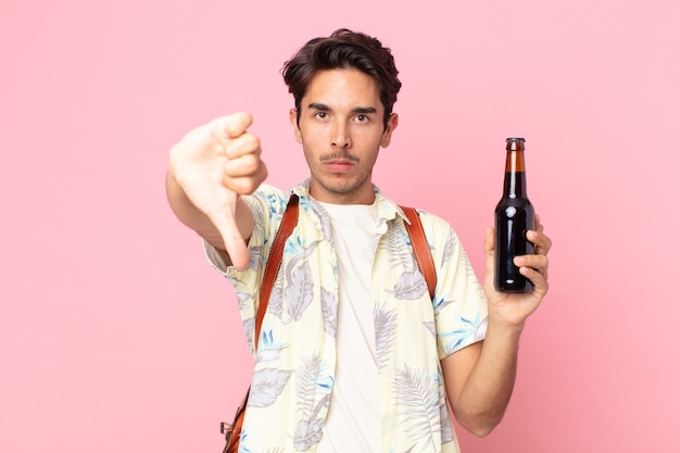 Joven hispano se siente cruzado, mostrando los pulgares hacia abajo y sosteniendo una botella de cerveza