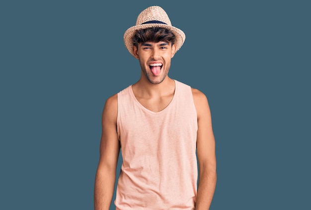 Joven hispano con sombrero de verano sacando la lengua feliz con expresión graciosa. concepto de emoción.