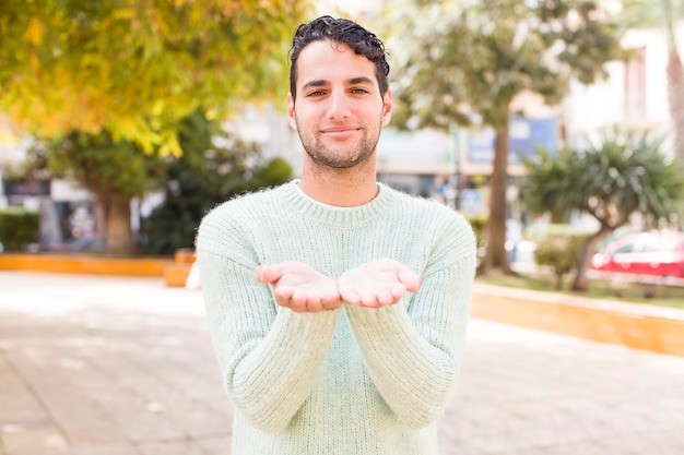 Foto joven hispano sonriendo alegremente con una mirada positiva y amigable ofreciendo y mostrando un obj