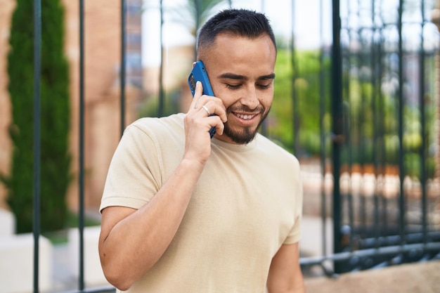 Joven hispano sonriendo confiado hablando por el teléfono inteligente en la calle