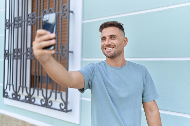 Joven hispano sonriendo confiado haciendo selfie por el teléfono inteligente en la calle